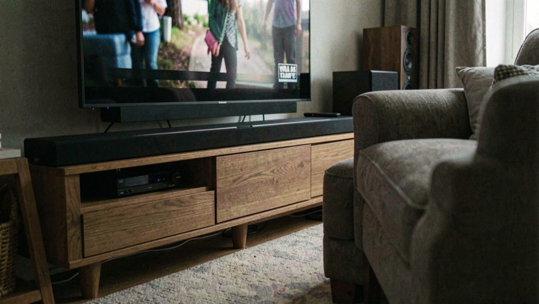 Soundbar nera allungata sotto una televisione accesa in un salotto in penombra, subwoofer appoggiato a pavimento vicino alla gamba del divano