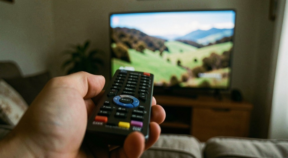 Telecomando tenuto in mano in primo piano sfocato davanti a uno schermo televisivo acceso su una scena di paesaggio, salotto in penombra